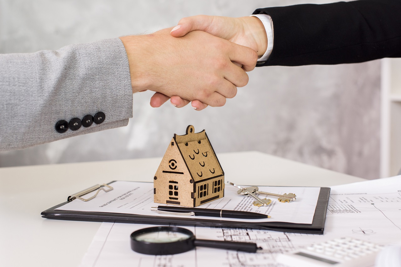 Two people in formal attire shaking hands over a wooden sculpture of a house