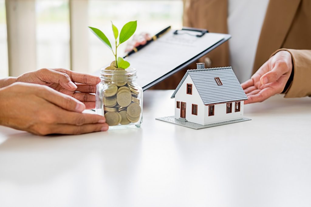 A hand offering a glass full of coins to a person with a miniature house symbolizing availing of a house loan