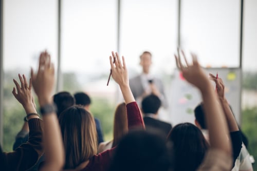 Multiple people have their hands raised. In the distance, blurred, is a man in front of these people, presumably delivering a sermon or lecture.