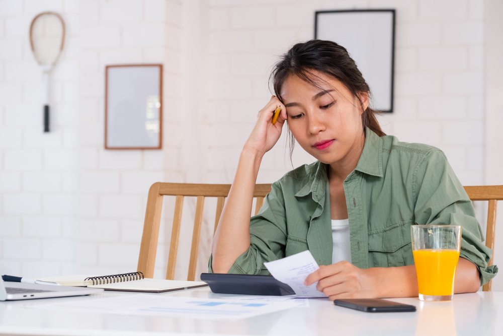 A young Asian woman sits at a table and looks worried as she looks at what looks to be a receipt she is holding in one hand. On the table in front of her are an open spiral notebook, a calculator, a smartphone, and a glass of an orange beverage. This is related to the blog's topic of the best reasons to get a personal loan.