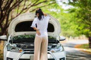 A young woman has her back to the camera. She has opened the hood of her car and seems frustrated. This is related to topic of how to save money on car repairs.