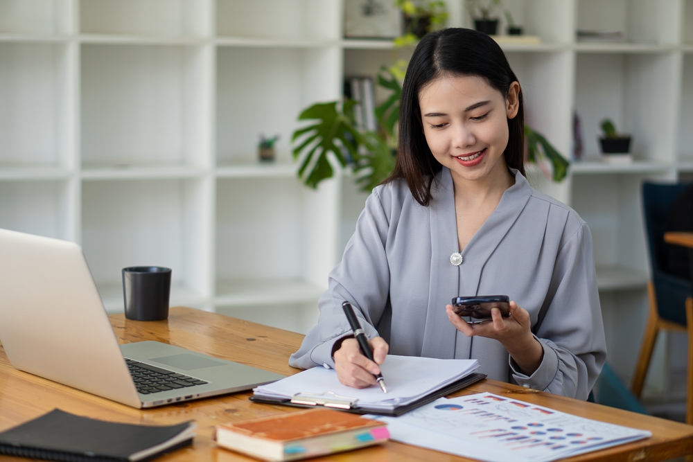A young Asian woman is sitting at her desk writing something down on a clipboard. In her other hand, she is holding her smartphone. On the desk in front of her are her open laptop, books, and sheets of paper showing what looks to be business projections. These are all related to the topic of what to consider before buying a franchise.