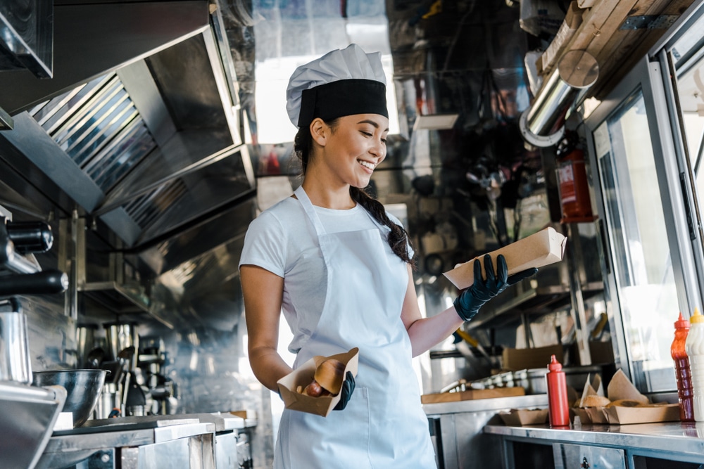 A young Asian woman is standing inside what appears to be the inside of a food truck. She is wearing an apron and holding two boxes of food in her hands. This is related to the concept of truck business ideas.